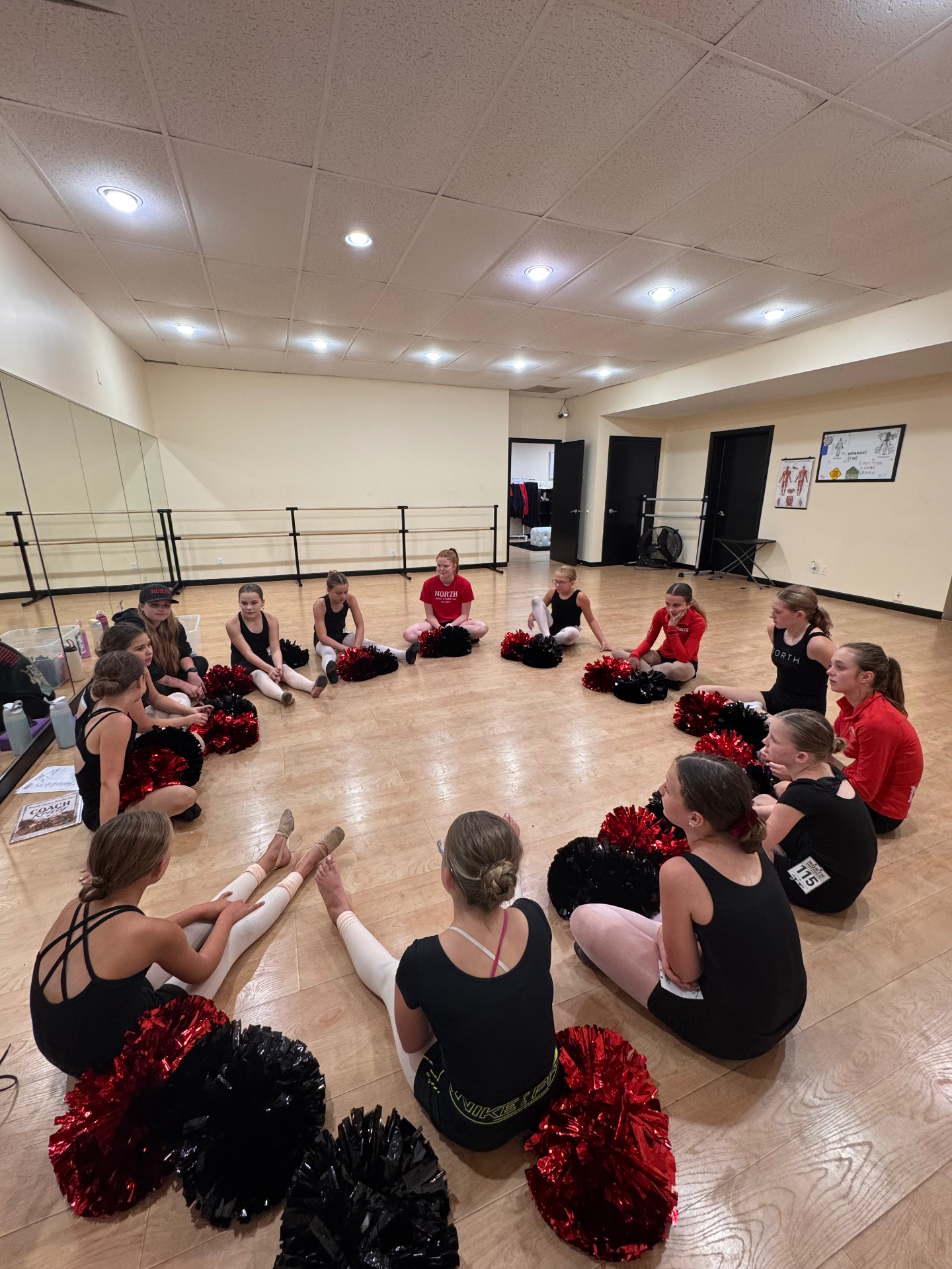Dancers sitting in a circle with red and black poms in the studio
